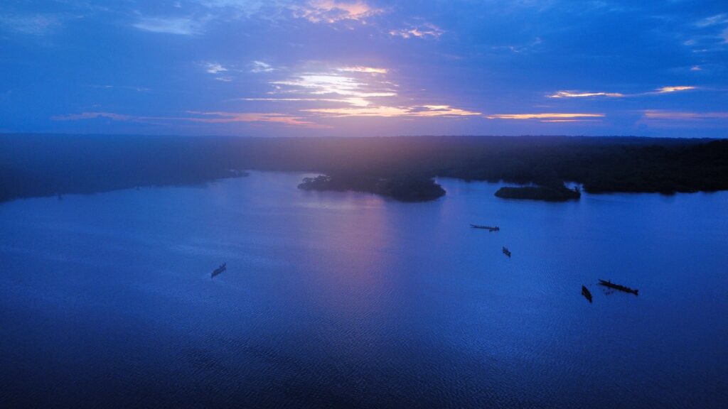 Vista aérea de la laguna al anochecer, con islas de bosque, varias canoas sobre el agua y la selva hasta el horizonte.