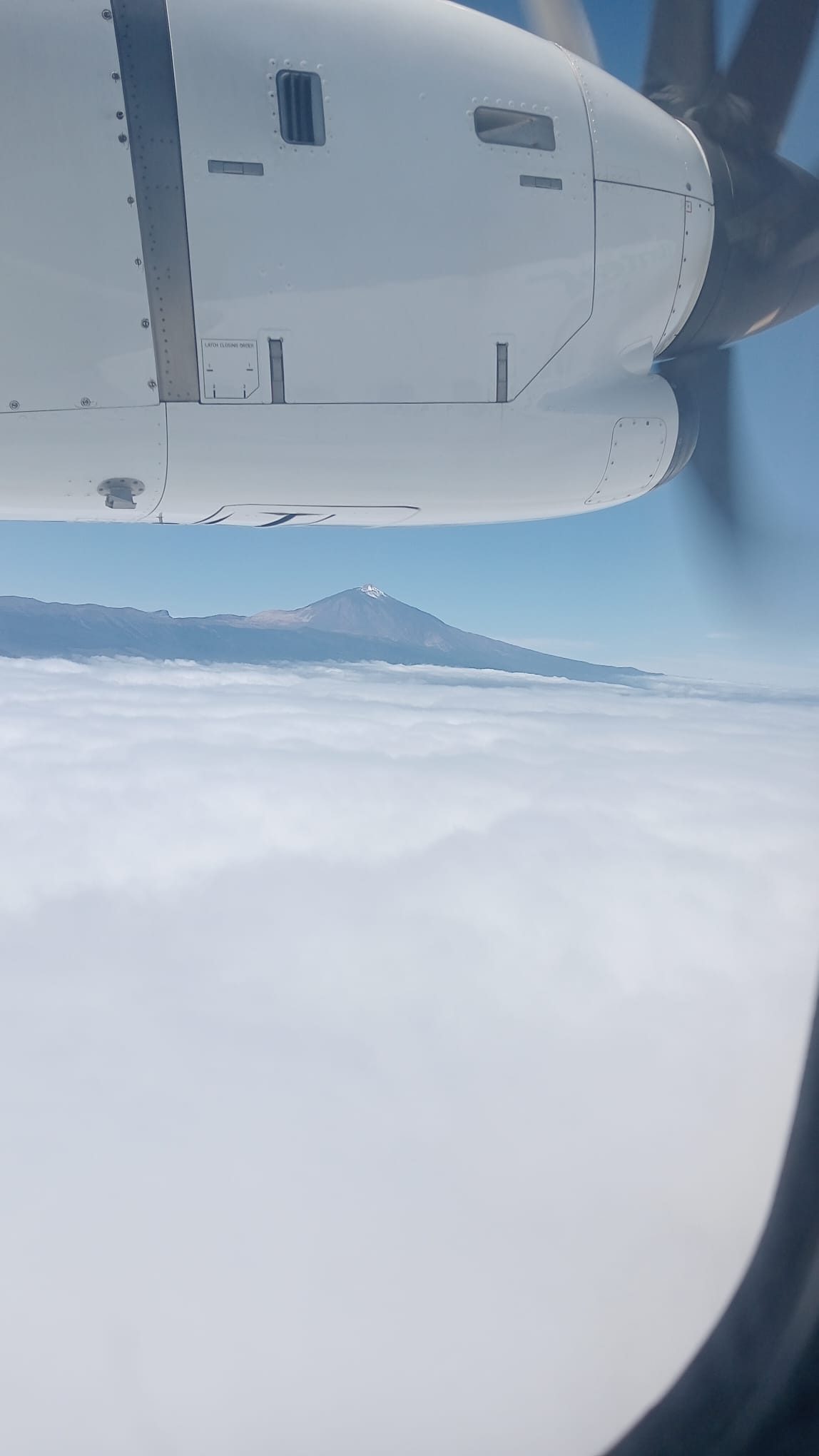 Ala y motor de un avión de hélice sobre un mar de nubes, con la cima de un volcán al fondo