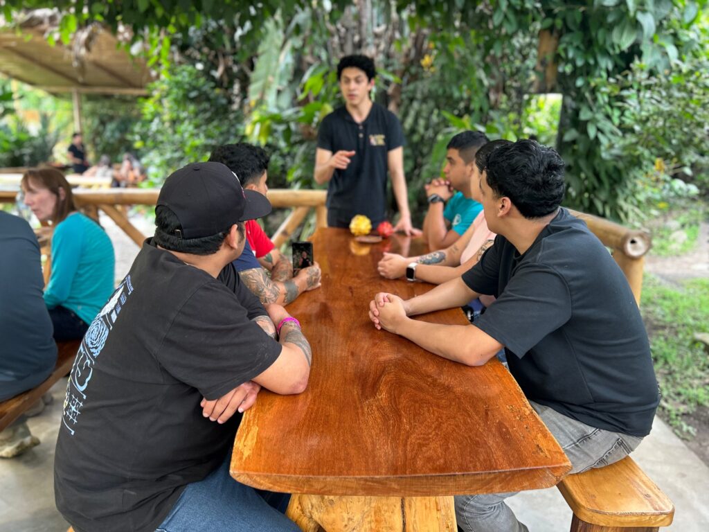 Guía explicando el proceso del cacao a un grupo de visitantes sentados en una mesa de madera