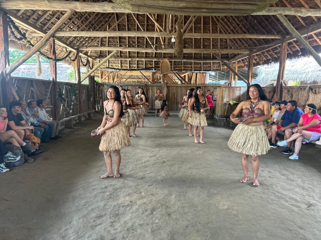 Mujeres de la comunidad bailan una danza tradicional frente a un grupo de visitantes en la casa comunal